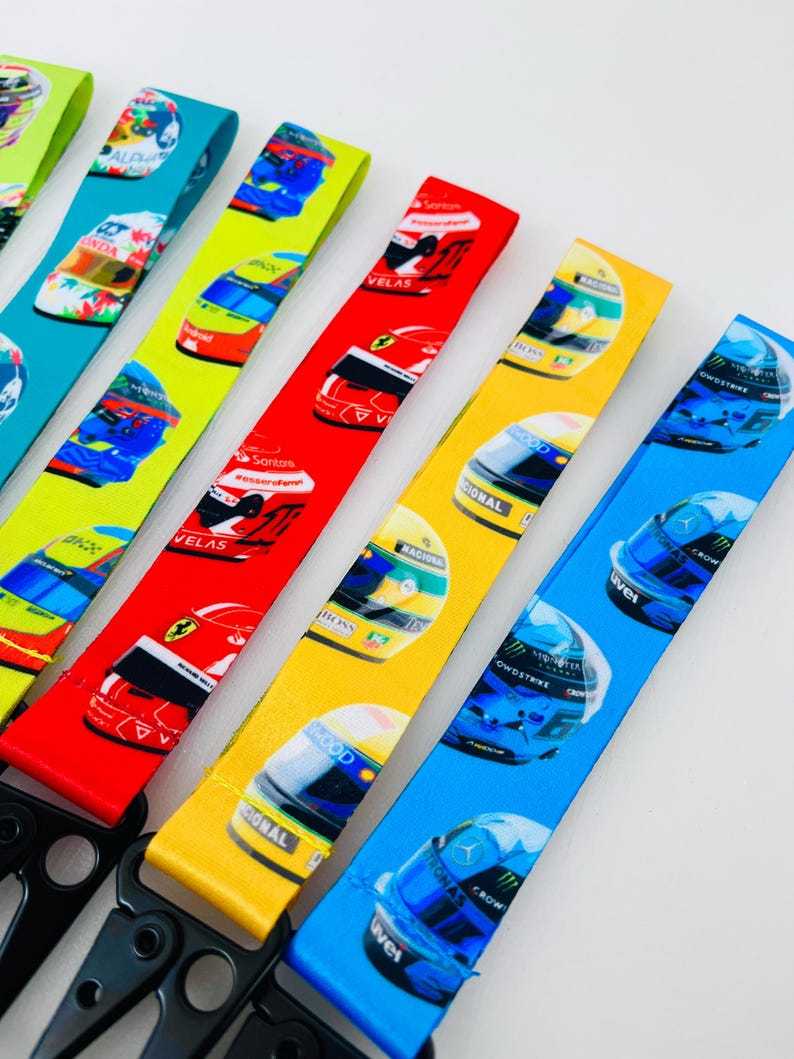 K&ouml;nnte beinhalten: F&uuml;nf bunte Lanyards mit verschiedenen Designs von Rennwagenhelmen. Die Lanyards sind aus weichem, gewebtem Stoff gefertigt und haben einen schwarzen Metallclip.