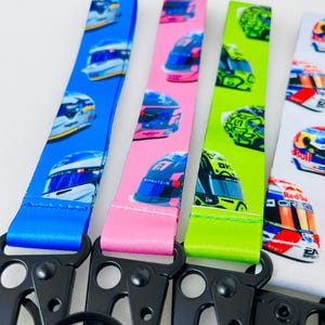 K&ouml;nnte beinhalten: Vier verschiedene farbige Lanyards mit einem schwarzen Metallclip und einem Ring. Jedes Lanyard zeigt ein sich wiederholendes Muster von Rennhelmen mit verschiedenen Sponsoren und Logos. Die Lanyards sind blau, rosa, gr&uuml;n und wei&szlig;.