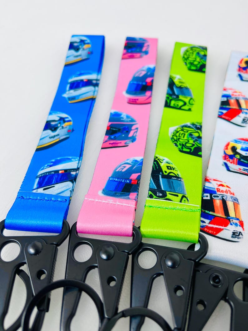 K&ouml;nnte beinhalten: Vier verschiedene farbige Lanyards mit einem schwarzen Metallclip und einem Ring. Jedes Lanyard zeigt ein sich wiederholendes Muster von Rennhelmen mit verschiedenen Sponsoren und Logos. Die Lanyards sind blau, rosa, gr&uuml;n und wei&szlig;.