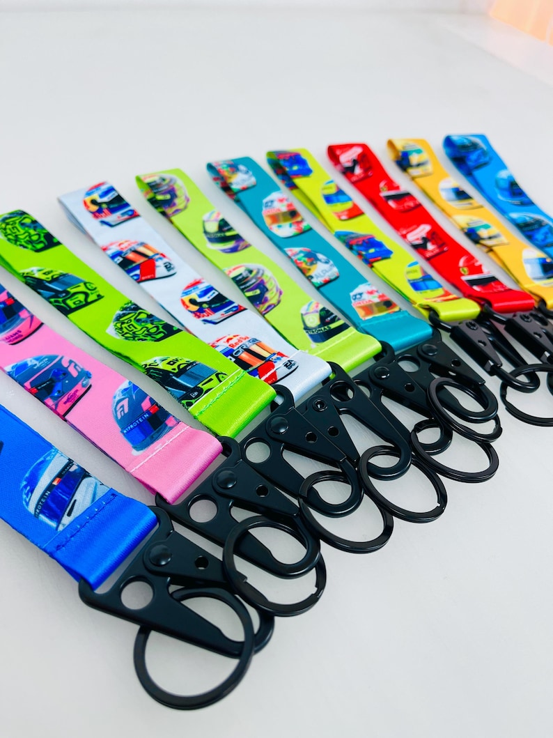 K&ouml;nnte beinhalten: Ein Set aus zehn bunten Lanyards mit einem Rennhelm-Muster und schwarzen Metall-Schl&uuml;sselanh&auml;ngern. Die Lanyards sind in verschiedenen Farben, darunter Gr&uuml;n, Blau, Rot, Gelb und Pink.