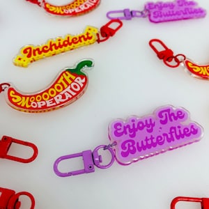 Könnte beinhalten: Sortiment an bunten Reißverschlussanhängern mit verschiedenen Sprüchen, darunter "Smooth Operator", "Incident" und "Enjoy The Butterflies". Die Reißverschlussanhänger sind in Form von Chilischoten und Schmetterlingen gestaltet.