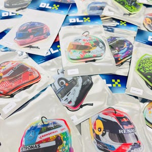 Puede incluir: Surtido de ambientadores de coche coloridos con forma de cascos de carreras. Los ambientadores presentan varios logotipos y patrocinadores de equipos de carreras, incluidos Red Bull, Renault, Petronas y Crowdstrike.
