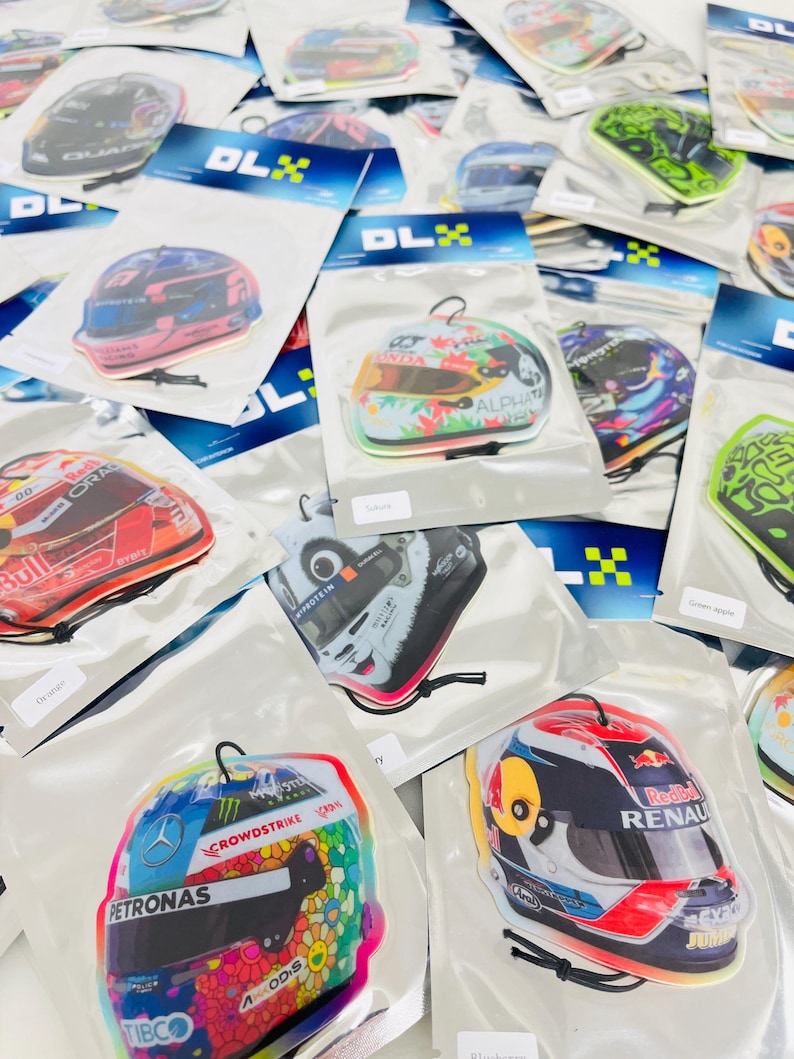Puede incluir: Surtido de ambientadores de coche coloridos con forma de cascos de carreras. Los ambientadores presentan varios logotipos y patrocinadores de equipos de carreras, incluidos Red Bull, Renault, Petronas y Crowdstrike.