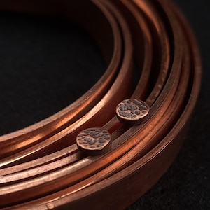 May include: Close-up of copper-colored metal earrings with a hammered texture. The earrings are circular and sit next to a series of curved copper bands. The background is dark.