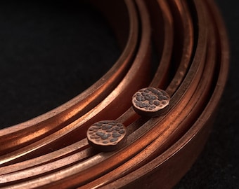 Puede incluir: Primer plano de pendientes de metal color cobre con textura martillada. Los pendientes son circulares y se encuentran junto a una serie de bandas de cobre curvadas. El fondo es oscuro.