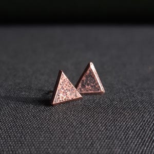 Könnte beinhalten: Ein Paar kupferfarbene Dreiecks-Ohrstecker. Die Ohrringe haben eine strukturierte Oberfläche und sind vor einem dunklen Hintergrund platziert. Das geometrische Design ist schlicht und modern.