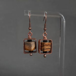 May include: A pair of copper earrings with a square frame and a brown and gold stone inside. The earrings are hanging from a clear stand.