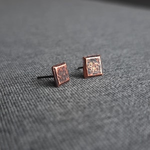 Könnte beinhalten: Ein Paar quadratische Ohrringe aus Kupfer mit gehämmerter Textur. Die Ohrringe haben einen schwarzen Stift und sind in einem schlichten und modernen Design gehalten.