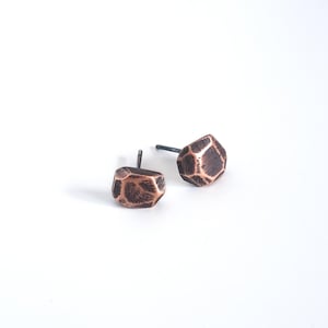 May include: A pair of copper earrings with a geometric, faceted design. The earrings have a dark, oxidized finish and are set on silver posts.