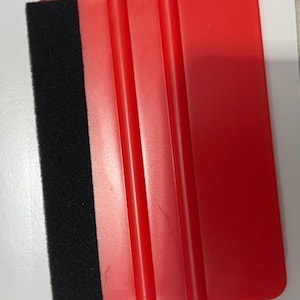 Puede incluir: Una espátula de plástico roja con un borde de espuma negra. La espátula tiene tres ranuras verticales en el mango. La herramienta está diseñada para aplicar vinilo, tinte para ventanas u otros materiales a una superficie.