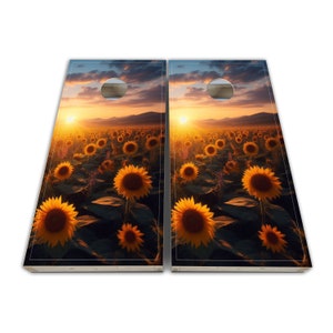 May include: Two wooden cornhole boards with a sunset over a field of sunflowers printed on them. The boards are set up for a game of cornhole.