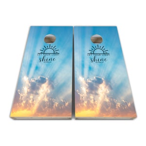 May include: A set of two wooden cornhole boards with a sky and clouds design. The boards have a sunrise graphic and the text "Let your light shine" with the scripture reference "Matthew 5:16".