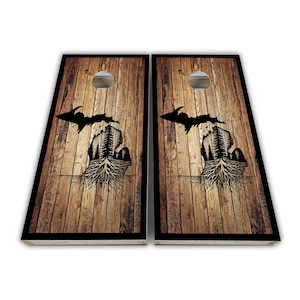 May include: Two wooden cornhole boards with a black outline. Each board features a wood grain background with a black silhouette of the state of Michigan, including a tree and root design. A silver-colored hole is at the top of each board.