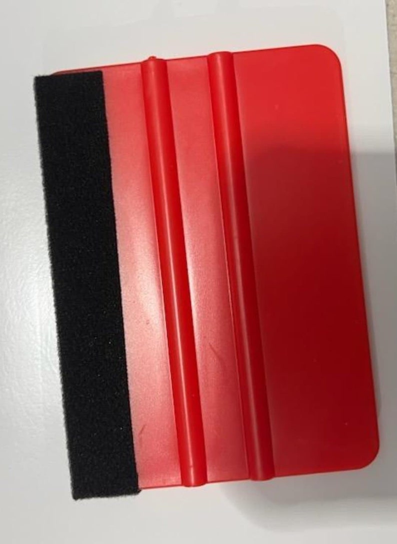 May include: A red plastic squeegee with a black felt edge. The squeegee has three raised ridges on the red surface.