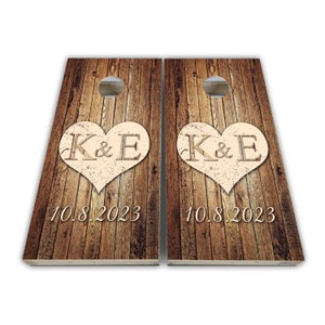 Könnte beinhalten: Zwei Holz-Cornhole-Bretter mit rustikalem Holzmaserungsmuster. Jedes Brett zeigt ein großes beigefarbenes Herz mit den Initialen "K & E" und dem Datum "10.8.2023" in cremefarbener Farbe.