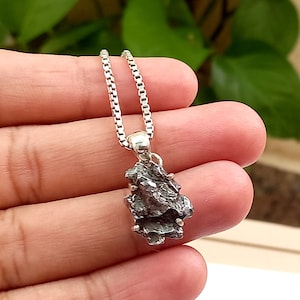 May include: A silver chain necklace with a pendant made of a rough, dark gray meteorite. The pendant is about 1 inch long and 1/2 inch wide.