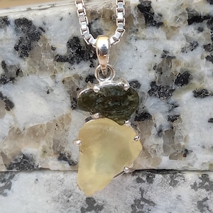 May include: A silver pendant necklace featuring two irregularly shaped gemstones. The top stone is a dark green color, and the bottom stone is a pale yellow. The pendant hangs from a delicate silver chain.