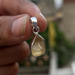 May include: A silver pendant with a raw, light yellow citrine gemstone. The citrine is irregularly shaped and set in a polished silver bezel. The pendant has a small loop for a chain.