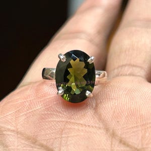 Può includere: Un anello d'argento con una grande pietra preziosa verde di forma ovale. L'anello è incastonato con quattro piccole griffe su ogni lato della pietra.
