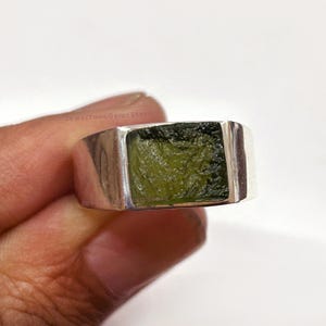 May include: A silver ring featuring a rectangular, olive-green gemstone. The stone has a textured surface and is set in a simple, polished silver band. The ring is shown against a white background.