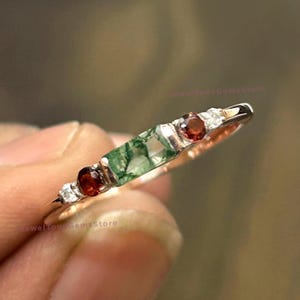 以下が含まれることがあります： 緑と白の瑪瑙の石と2つの赤いガーネットの石が付いた銀のリング。リングは銀のバンドにセットされています。