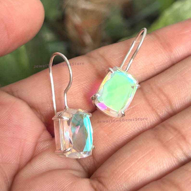 May include: A pair of silver drop earrings featuring rectangular, faceted iridescent stones that display shifting colors of green, pink, and white. The earrings have simple silver hooks and the text JewelToneGemsStore is visible on the stones.