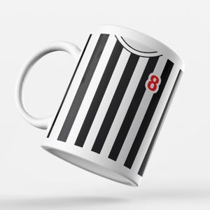 Puede incluir: Taza de cer&aacute;mica blanca con rayas verticales negras, un n&uacute;mero 8 rojo y un asa blanca. La taza tiene un dise&ntilde;o que se asemeja a una camiseta deportiva. La taza est&aacute; ligeramente inclinada hacia la izquierda.