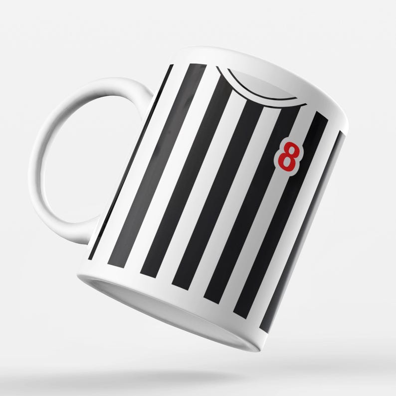 Puede incluir: Taza de cer&aacute;mica blanca con rayas verticales negras, un n&uacute;mero 8 rojo y un asa blanca. La taza tiene un dise&ntilde;o que se asemeja a una camiseta deportiva. La taza est&aacute; ligeramente inclinada hacia la izquierda.