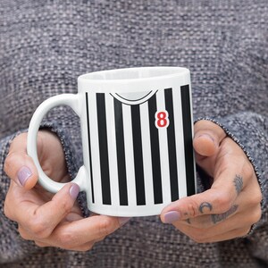Puede incluir: Taza de cer&aacute;mica blanca con un dise&ntilde;o a rayas blancas y negras, con el n&uacute;mero 8 en rojo. La taza tiene un asa curva. El dise&ntilde;o se asemeja a una camiseta deportiva.