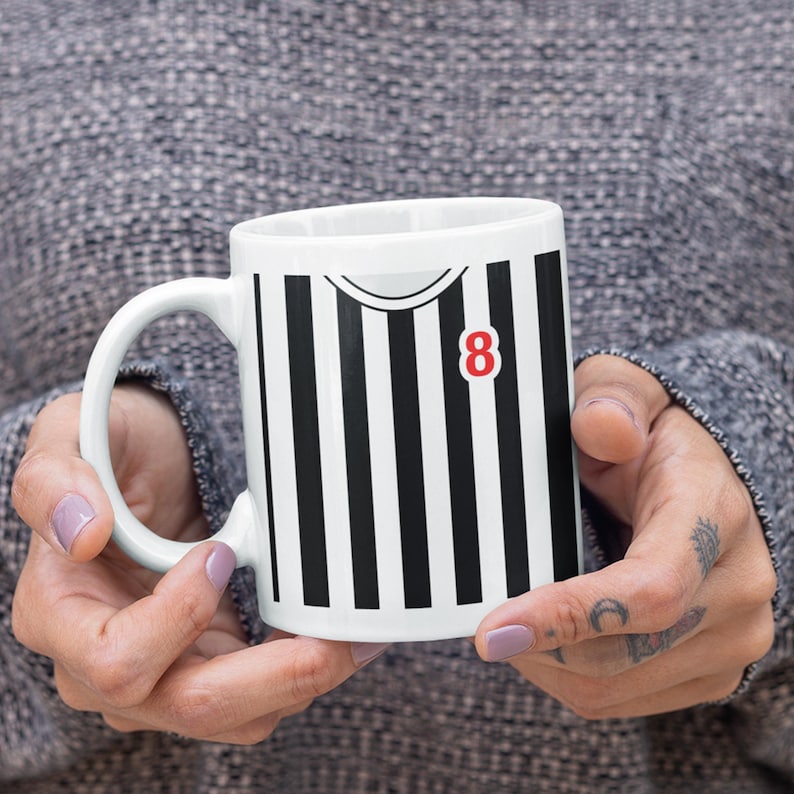 Puede incluir: Taza de cer&aacute;mica blanca con un dise&ntilde;o a rayas blancas y negras, con el n&uacute;mero 8 en rojo. La taza tiene un asa curva. El dise&ntilde;o se asemeja a una camiseta deportiva.