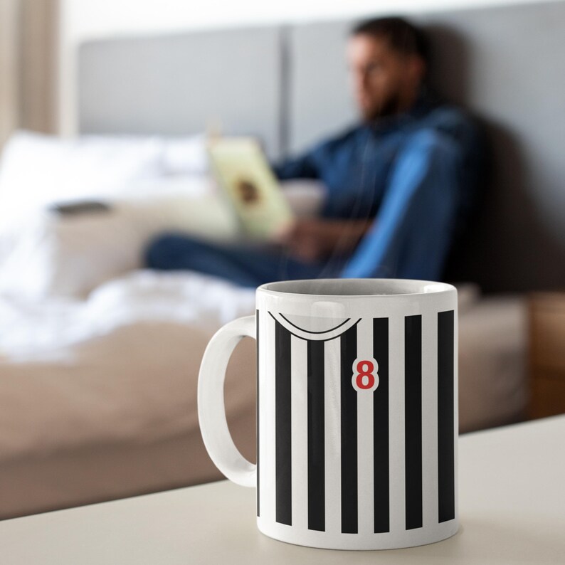 Puede incluir: Taza de cer&aacute;mica blanca con un dise&ntilde;o a rayas blancas y negras, con el n&uacute;mero 8 en rojo. La taza tiene un asa curva y un dise&ntilde;o de cuello de camisa. La taza est&aacute; sobre una superficie blanca.