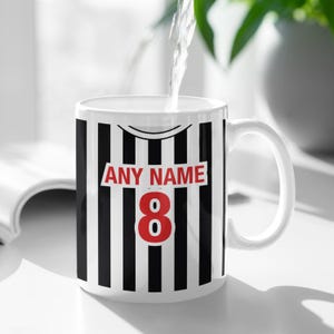 Puede incluir: Taza de cer&aacute;mica blanca con rayas verticales negras, el n&uacute;mero 8 en rojo y el texto "ANY NAME". La taza tiene un asa curva y se est&aacute; llenando de agua. El dise&ntilde;o se asemeja a una camiseta deportiva.