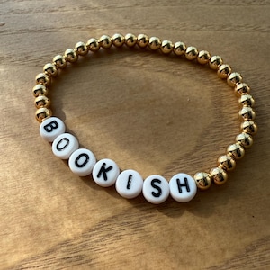 May include: Gold beaded bracelet with white letter beads spelling "BOOKISH". The bracelet features small, round gold beads and white beads with black letters. The bracelet is displayed on a wooden surface.