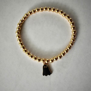 Może przedstawiać: Złota bransoletka z koralików z czarnym urokiem ducha. Bransoletka wykonana jest z małych, okrągłych koralików i ma prosty, elegancki design.