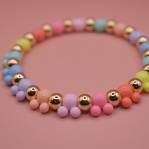 May include: A rainbow coloured beaded bracelet with gold accents. The beads are arranged in a gradient pattern, starting with yellow and ending with pink.
