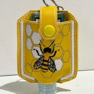 May include: Yellow hand sanitiser holder with a bee and honeycomb design. The holder has a white trim and a yellow button closure. It has a silver key ring.
