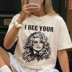 May include: An ivory short-sleeve t-shirt featuring a black graphic illustration of a woman with voluminous hair and the text I BEG YOUR PARDON printed in a bold, stylized font across the chest. The shirt has a crew neckline and a relaxed fit.