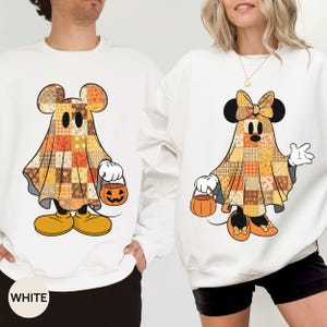 May include: Two white crewneck sweatshirts featuring Halloween-themed graphics. The left sweatshirt displays a ghost character with a patchwork pattern in orange, yellow, and brown, holding a pumpkin bucket. The right sweatshirt shows a similar ghost character with a bow, also holding a pumpkin bucket.