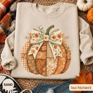 May include: A sand-coloured crewneck sweatshirt featuring a large central graphic of a pumpkin. The pumpkin is designed with a patchwork pattern of various autumn-themed fabrics, including gingham and floral prints, topped with a large floral bow.