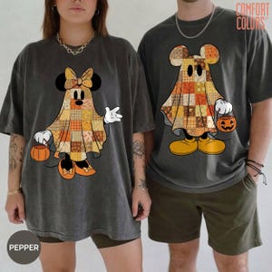 Puede incluir: Dos camisetas gris carbón con estampados gráficos de fantasmas con patrones de patchwork en tonos naranja, amarillo y marrón. La camiseta izquierda muestra un fantasma con un lazo y la derecha un fantasma con un cubo de calabaza.
