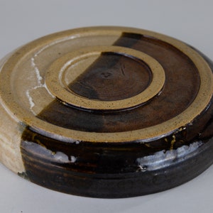 May include: A round ceramic plate with a brown and white glaze. The plate has a raised rim and a slightly uneven surface.