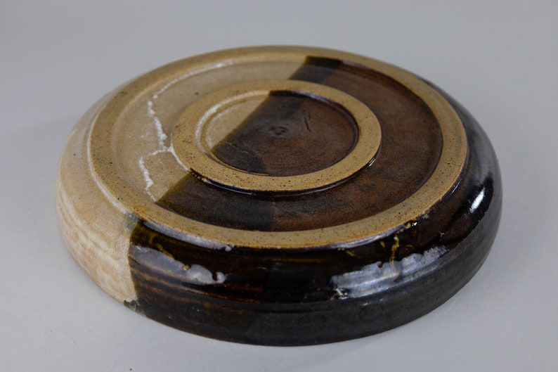 May include: A round ceramic plate with a brown and white glaze. The plate has a raised rim and a slightly uneven surface.