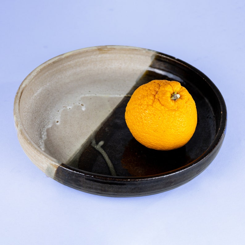 May include: A single orange citrus fruit sits on a black and white ceramic plate. The plate has a brown glaze and a white glaze, creating a two-toned effect.