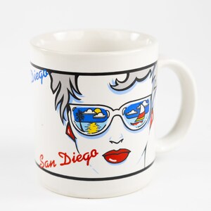 May include: White ceramic mug with a colorful illustration of a woman wearing sunglasses. The sunglasses reflect a beach scene with a palm tree, sun, and clouds. The text "San Diego" is written in red on the mug.