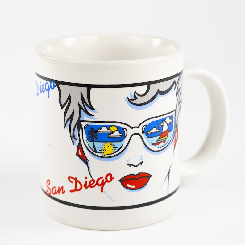 May include: White ceramic mug with a colorful illustration of a woman wearing sunglasses. The sunglasses reflect a beach scene with a palm tree, sun, and clouds. The text "San Diego" is written in red on the mug.