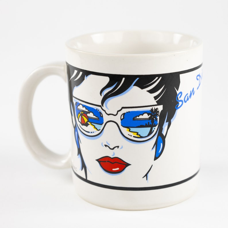 May include: White ceramic mug with a colorful illustration of a woman wearing sunglasses. The sunglasses reflect a beach scene with an umbrella, palm trees, and blue sky. The text "San Diego" is printed on the mug.