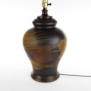 May include: A brown ceramic table lamp with a gold and brown abstract design. The lamp has a wooden base and a black cord.