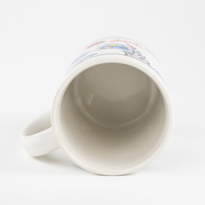 May include: White ceramic mug with a blue and red design. The mug has a handle and is empty.