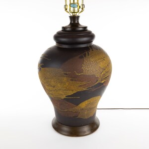 May include: A brown ceramic table lamp with a gold and red abstract design. The lamp has a round base and a cylindrical shade.