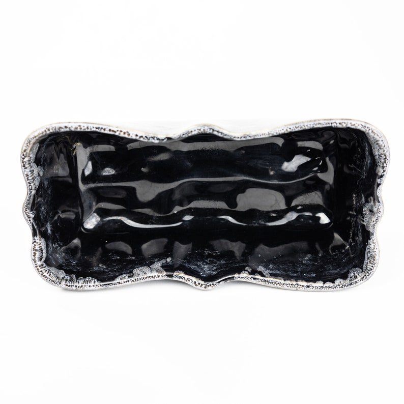 May include: A black ceramic rectangular serving dish with a wavy, silver-colored rim. The dish is empty and has a glossy finish.